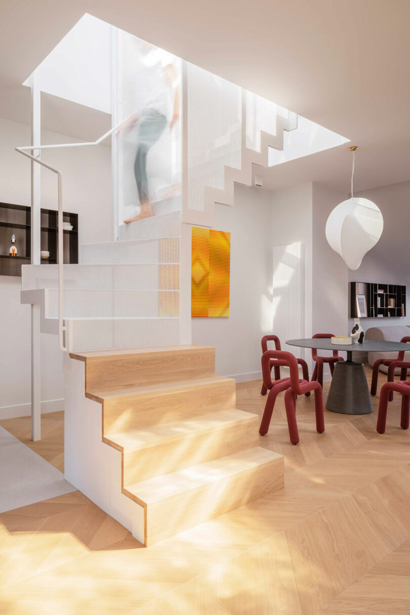 A modern interior features a wooden staircase with a white metal upper section, geometric furniture, abstract art, and sunlight streaming through a skylight.