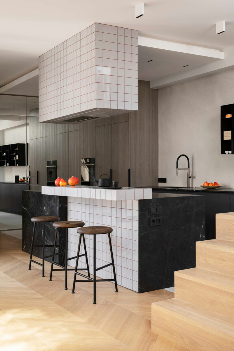 Modern kitchen with a black marble island, white square tile accents, two bar stools, built-in appliances, and wooden flooring; oranges sit on the counter.