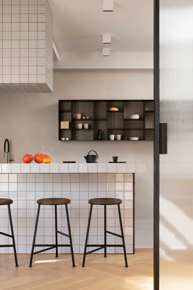 Modern kitchen with a white tiled island, three bar stools, a black faucet, teapot, cups, and fruits on the counter; open shelving with dishes in the background.