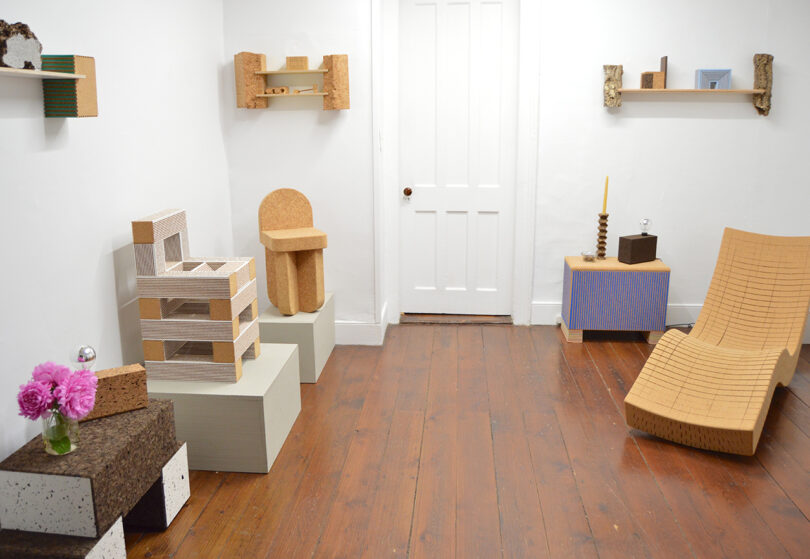 A minimalist room with geometric cork furniture, white walls, wooden floor, shelves with small objects, and a vase of pink flowers on a side table.