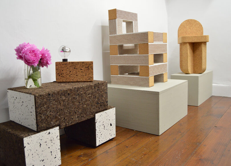 Geometric sculptures made of cork, cardboard, and foam are displayed on painted wooden plinths; pink flowers and a small reflective sphere sit atop one piece.