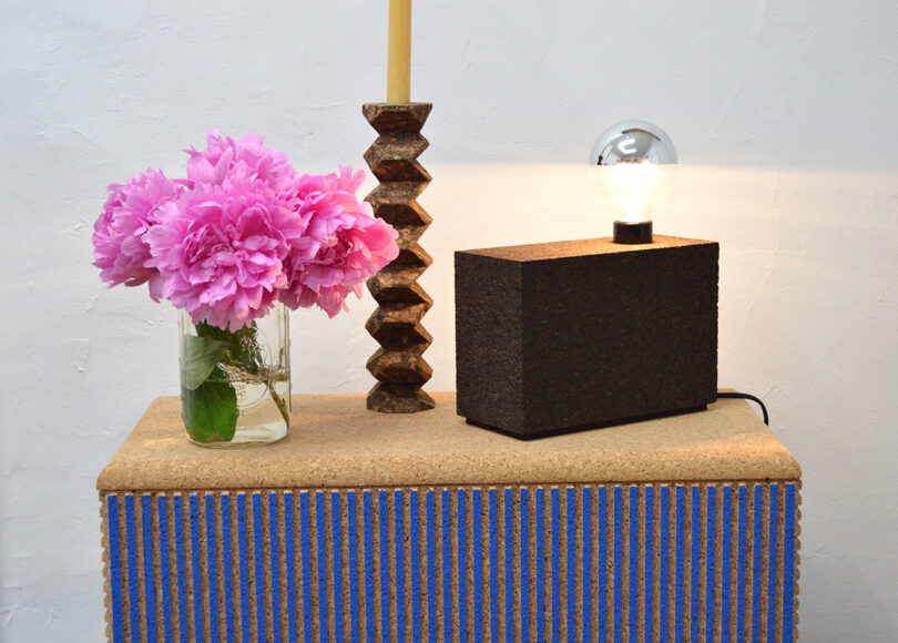A glass jar of pink flowers, a tall twisted brown candlestick with a yellow candle, and a rectangular lamp with an exposed bulb sit on a striped blue and beige table.