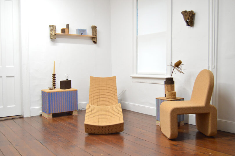 Minimalist room with wooden floor, three modern cork chairs, two box-shaped tables, wall-mounted shelf with decor, and white walls with small art pieces.