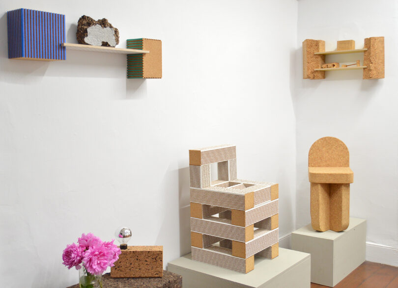A minimalist room with sculptural furniture and shelves made of cork and wood, a vase of pink flowers, and geometric art objects displayed on pedestals and walls.
