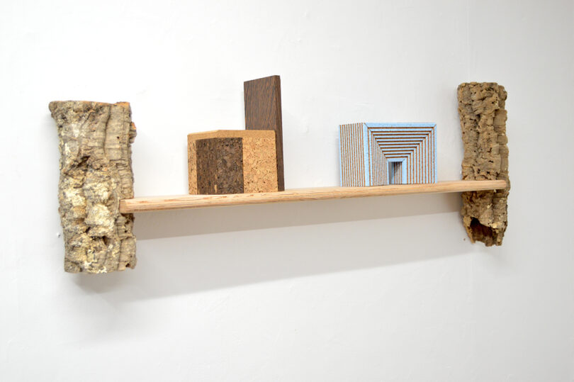 A wooden shelf supported by two pieces of bark holds three geometric blocks made of different materials against a white wall.