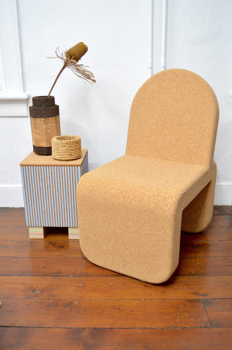 A minimalist cork chair sits next to a small side table with a striped front, holding decorative vases/