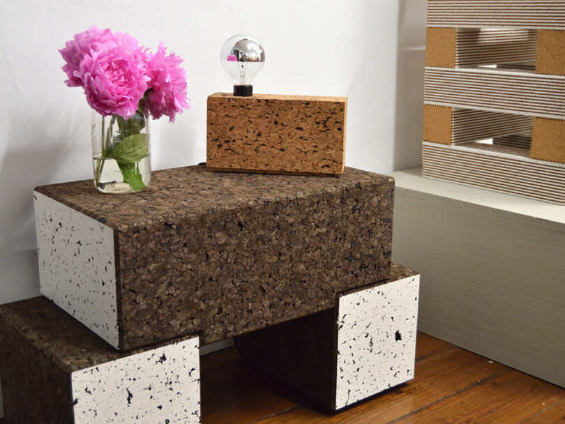 A glass jar with pink flowers, a cork block with a small bulb, and stacked rectangular blocks made of cork and white speckled material sit on a wooden floor next to a cardboard structure.