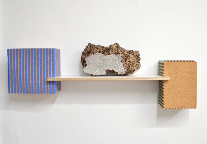 A wooden shelf mounted on a white wall holds a textured stone between two rectangular blocks with colorful horizontal stripes and cork surfaces.