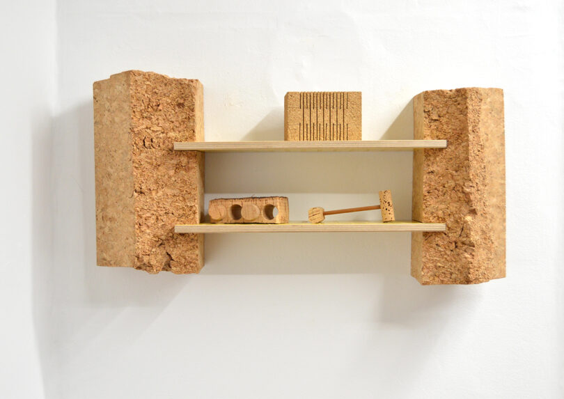 Wall-mounted wooden shelf with thick cork blocks at each end, holding small cork and wood objects arranged on two shelves against a white wall.