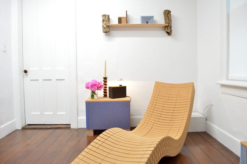 A minimalist room with a wooden lounge chair, a small side table holding a vase of pink flowers, a candle, and a box, and a shelf with decor on a white wall.