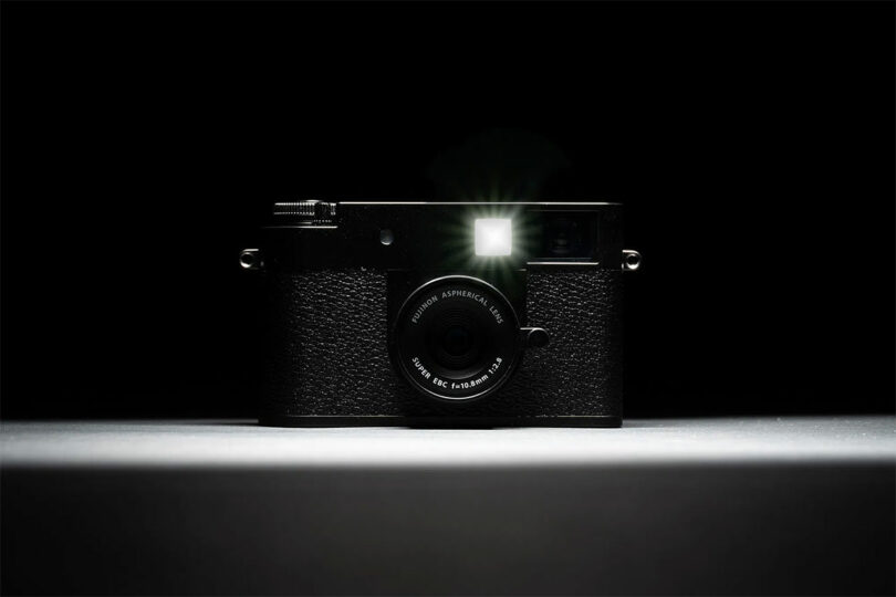 A Fujifilm black camera is positioned on a surface in a dark setting, facing forward with its flash illuminated.