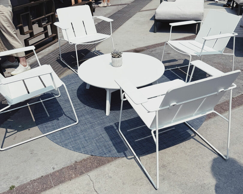 Four white metal chairs surround a round white table with a small potted plant, set on a blue outdoor rug on a concrete patio—perfect for relaxing or gathering after exploring Fulton Market DesignDays.