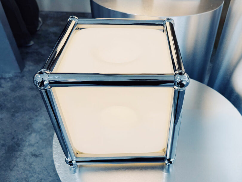 A cube-shaped light fixture with frosted white sides and shiny chrome corner supports, inspired by Fulton Market DesignDays, placed on a metallic circular surface.