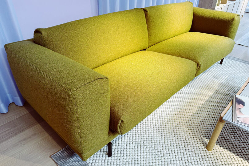 A modern olive green sofa with wide armrests sits on a textured light-colored rug next to a wooden coffee table with magazines, echoing the contemporary style seen at Fulton Market DesignDays.