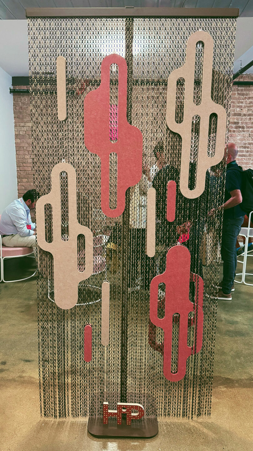 A decorative room divider with pink and beige geometric shapes hangs in front of a group of people at Fulton Market DesignDays, set within an indoor space featuring brick walls.