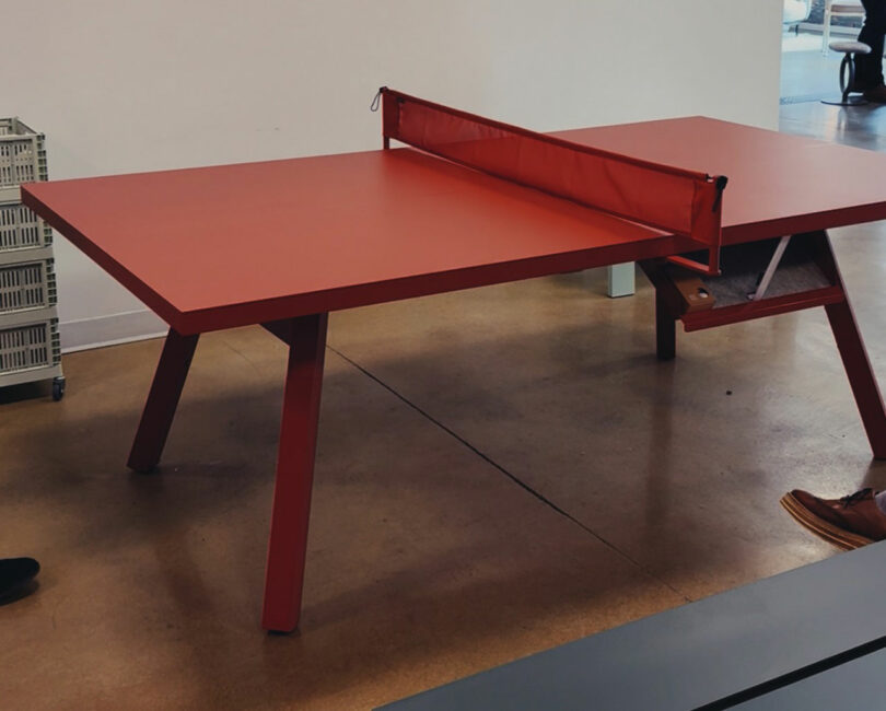Red ping pong table with a matching red net, set up indoors on a brown floor; partial view of people’s shoes visible near the table—capturing the vibrant energy of Fulton Market DesignDays.