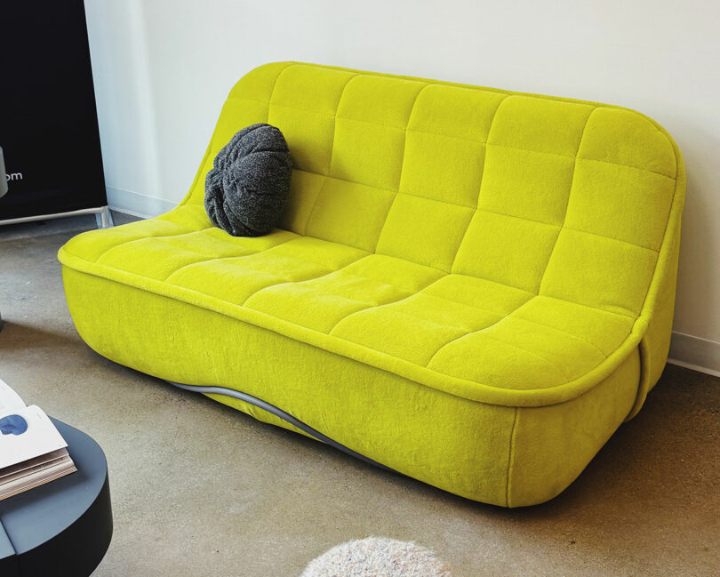 A bright yellow, modern sofa with a quilted pattern is placed against a white wall at Fulton Market DesignDays, featuring a round gray pillow on one side.