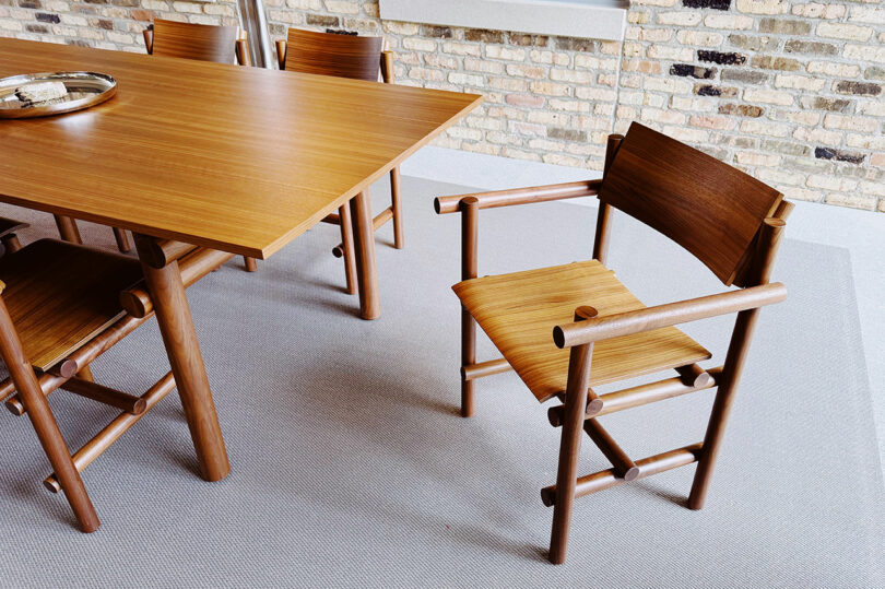 A wooden dining table with matching wooden chairs sits on a light textured rug against an exposed brick wall, evoking the contemporary charm of Fulton Market DesignDays.