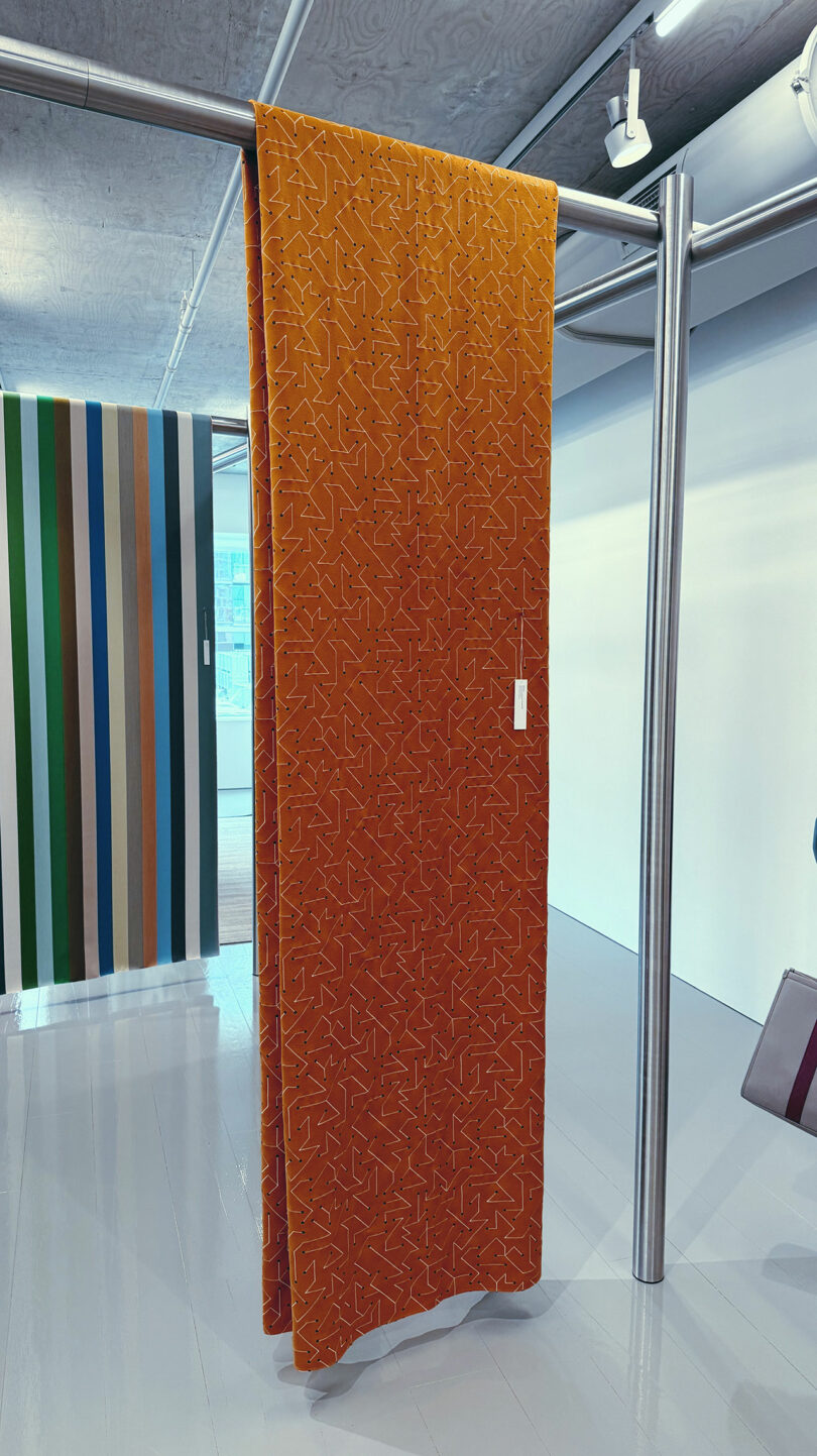 A large orange fabric with a subtle geometric pattern hangs on a metal rail in a modern showroom at Fulton Market DesignDays; striped fabrics are visible in the background.