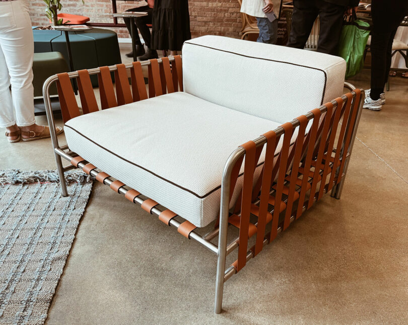Modern lounge chair with a metal frame, brown leather strap sides, and white cushioned seat and backrest, showcased on a concrete floor during Fulton Market DesignDays in a stylish showroom setting.