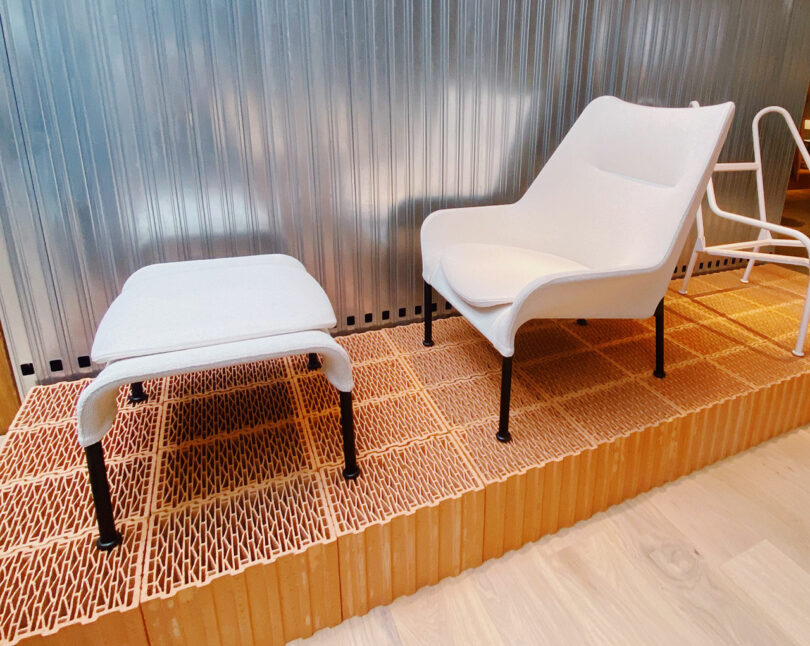 A modern white lounge chair with matching ottoman sits on a raised platform with a textured surface, next to a corrugated metal wall, reflecting the contemporary vibe of Fulton Market DesignDays.