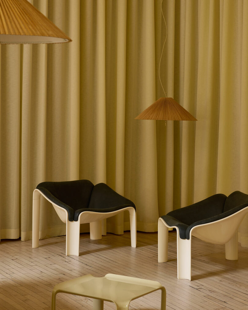 Two modern F300 Pierre Paulin chairs with black seats and white legs sit near beige curtains, paired with a matching side table and a brown pendant lamp above.