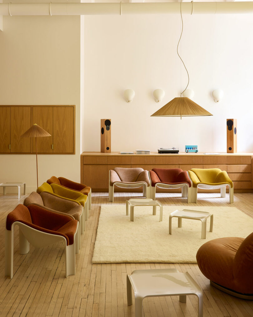 A modern lounge featuring F300 Pierre Paulin curved chairs in warm colors, low white tables, a cream rug, wood cabinetry, speakers, and a pendant light in a minimalist setting.