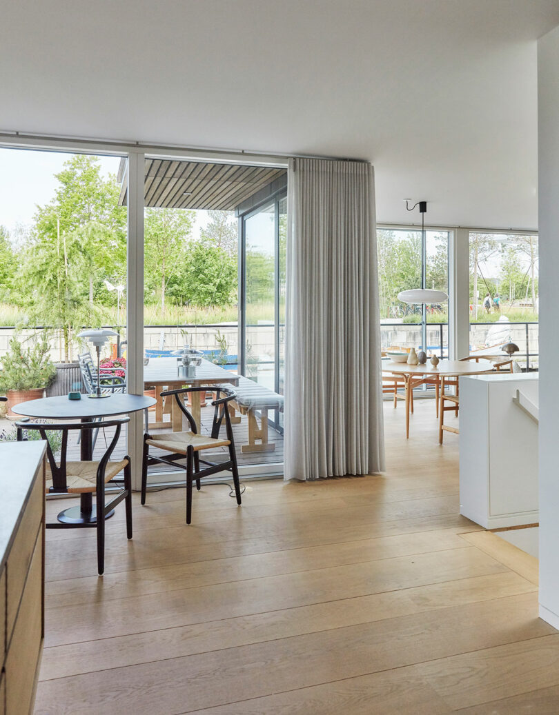 Modern dining area with wooden floors, glass doors, and outdoor seating visible through large windows. Simple furniture and neutral decor create a minimalist, airy atmosphere.