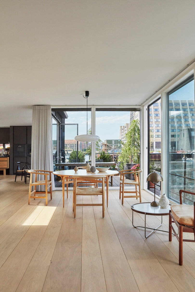 Modern dining area with wooden furniture, large windows, and views of water and city buildings. Light wood floor and minimalist decor create a bright, airy atmosphere.