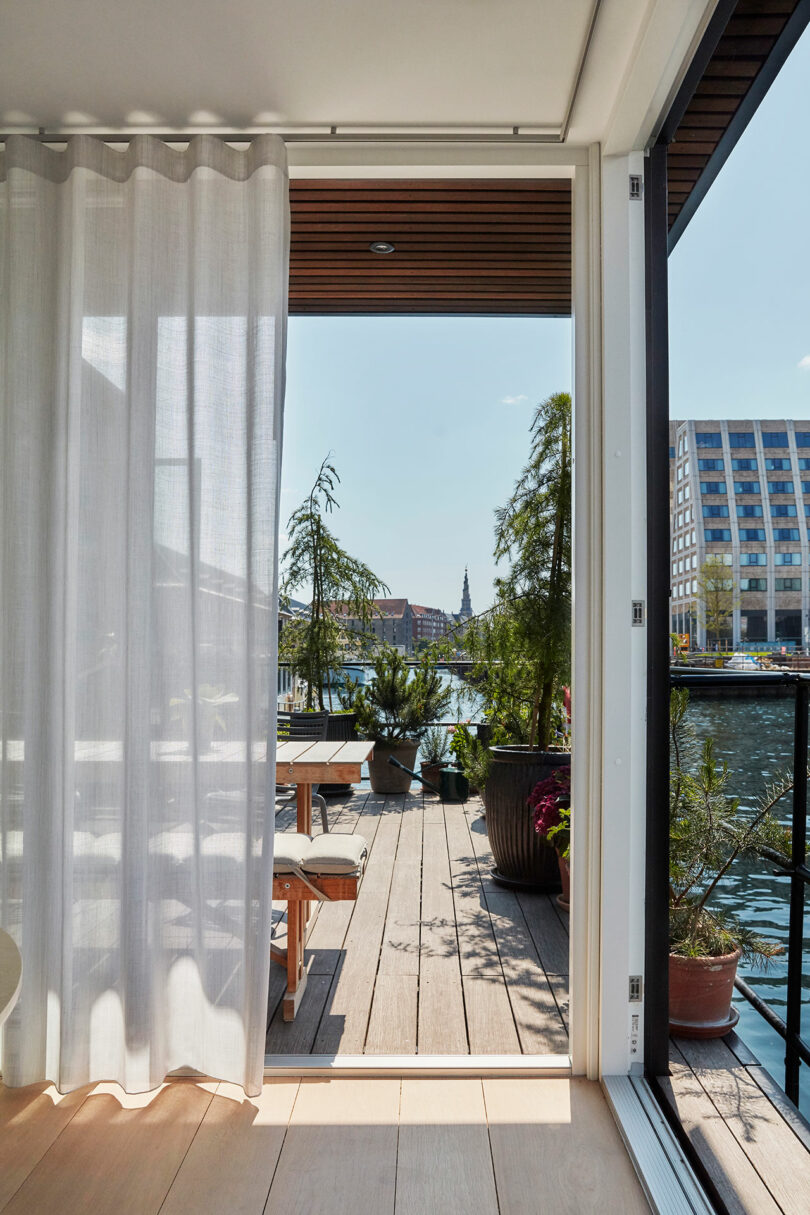 View from inside a room through an open sliding door to a wooden deck with potted plants, outdoor seating, and a waterfront cityscape in the background.
