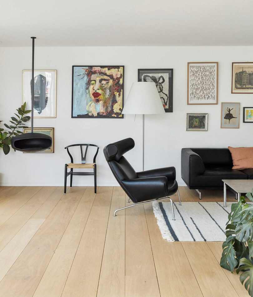 Modern living room with light wood floors, black leather chairs, a white floor lamp, a striped rug, and a gallery wall of various framed artworks above a black sofa.