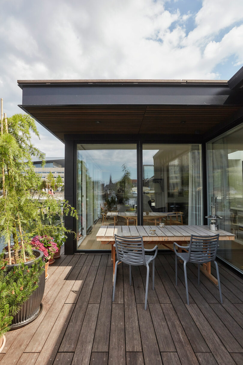 A modern patio with wooden decking, potted plants, a table and chairs, and large glass doors leading to an indoor dining area.