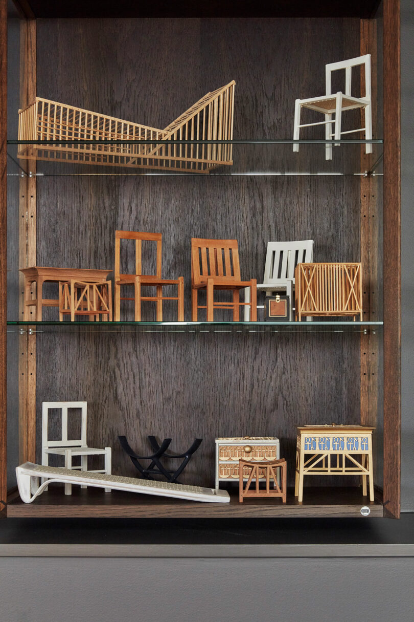 A display cabinet with shelves holding miniature models of chairs, benches, and furniture pieces in various designs and materials.