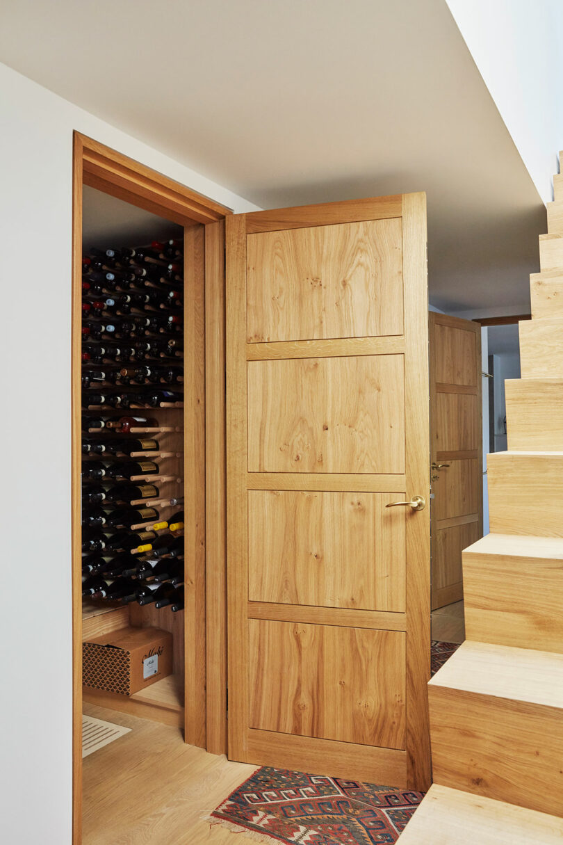 A wooden door beside a staircase opens to reveal a wine storage room with bottles organized on shelves.