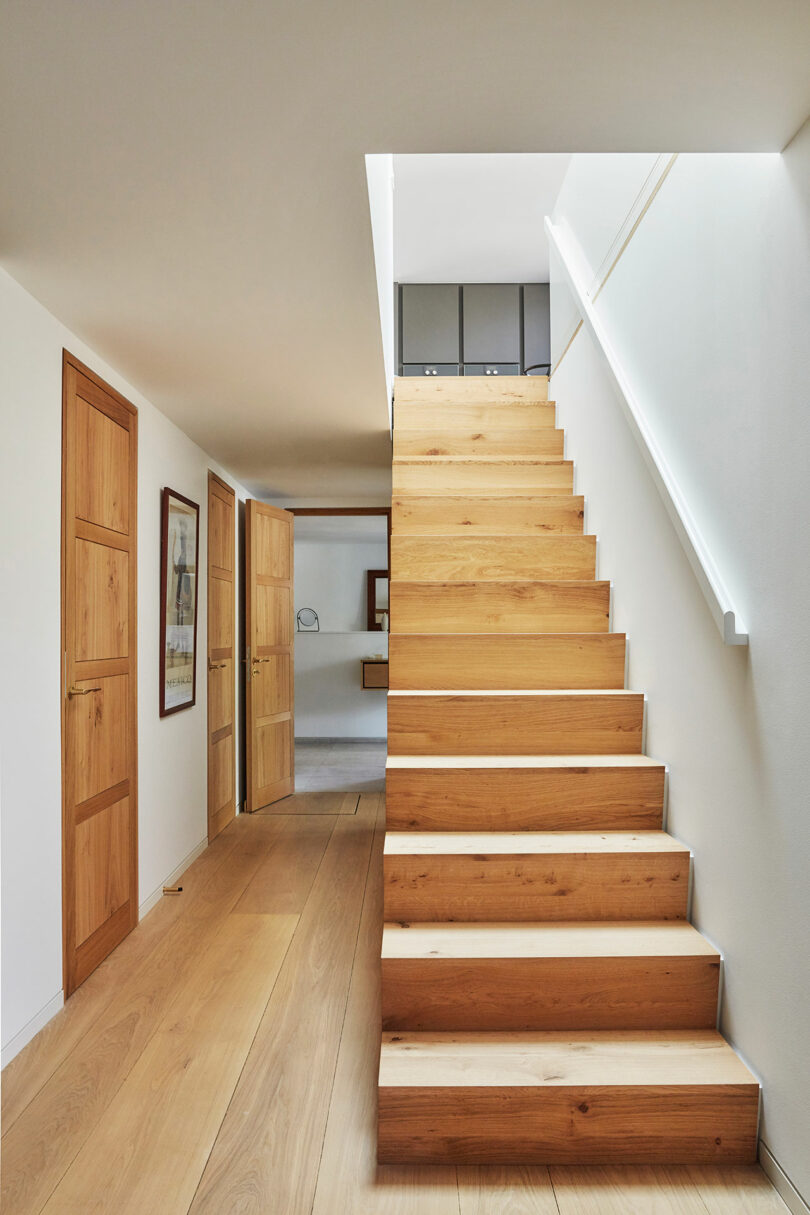 Wooden staircase with white walls, wooden doors on the left, and a hallway leading to another room in a modern, minimalist interior.