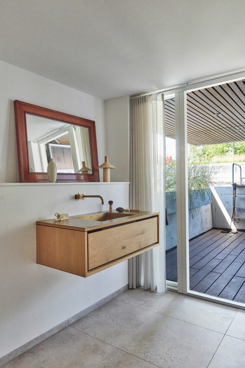 Minimalist bathroom with a floating wooden sink, wall-mounted faucet, framed mirror, ceramic decor, and a glass door leading to an outdoor deck.