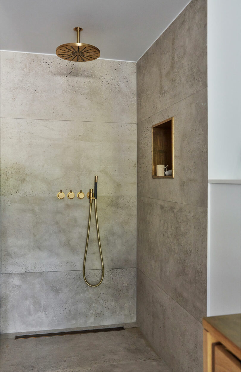 Minimalist shower with concrete walls, brass rain showerhead, three brass controls, a handheld shower, and a built-in niche holding soap and a small item.