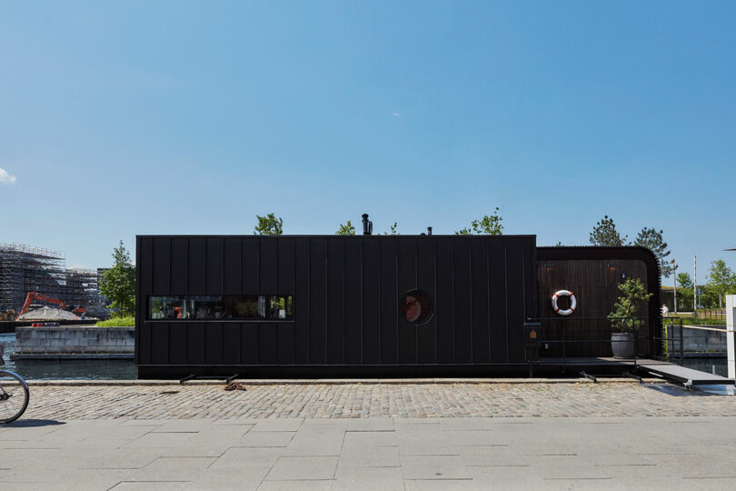 A modern black houseboat with rectangular windows and a round porthole is docked along a cobblestone path by the water under a clear blue sky.