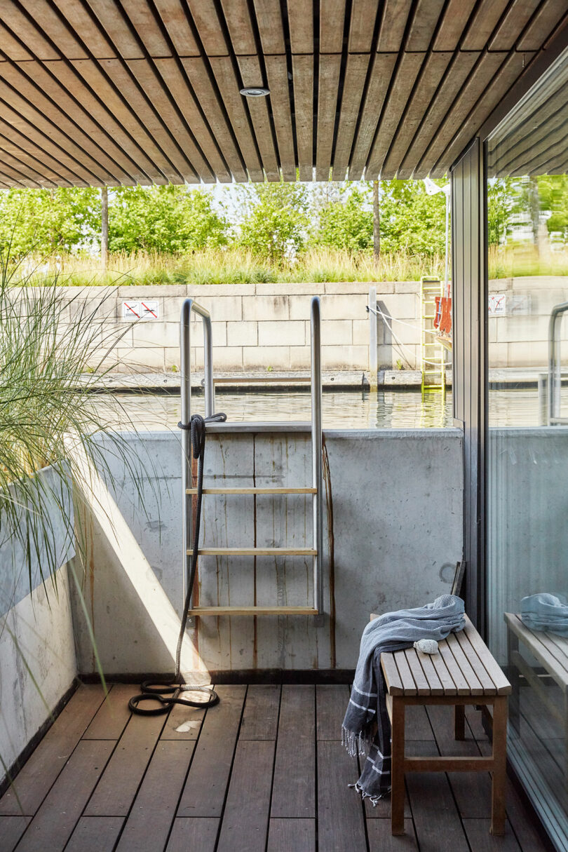 A small wooden deck with a bench and towel, a ladder leading into the water, and a concrete wall with a glass door and greenery visible outside.