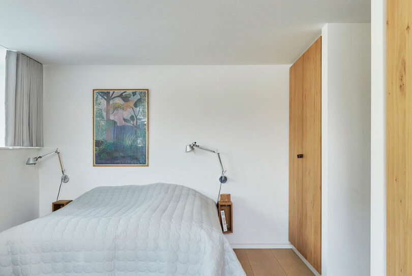 A minimalist bedroom with a neatly made bed, two wall-mounted reading lamps, a framed painting above the bed, and a wooden wardrobe.
