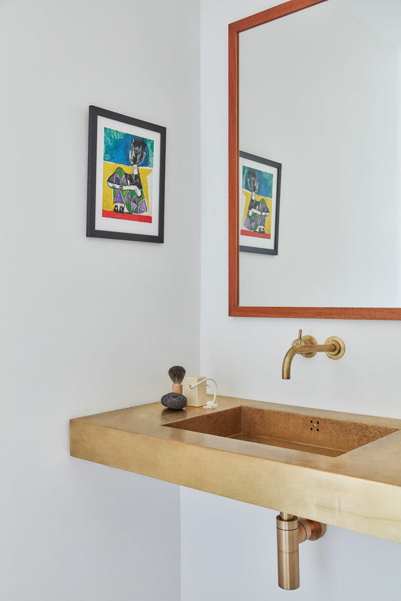 Minimalist bathroom with a gold sink and faucet, wall-mounted mirror with a wooden frame, and a colorful framed artwork on a white wall.