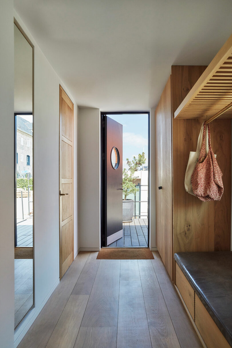 A modern entryway with wooden flooring, a built-in bench, a hanging bag, a door with a round window open to the outside, and a tall wall mirror.