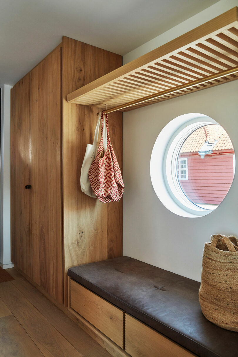 A wooden entryway with built-in storage, a leather bench, two hanging bags, a circular window, and a woven basket on the floor.