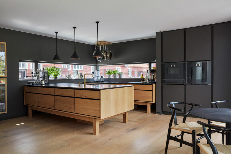 Modern kitchen with wood cabinets, black countertops, pendant lights, built-in ovens, and large windows; potted plants sit on the windowsill and natural light fills the space.