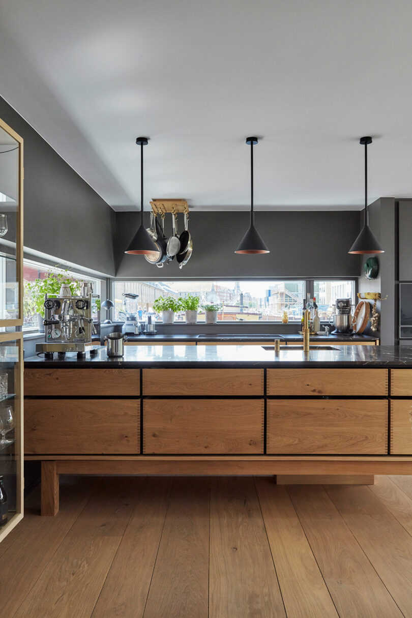 Modern kitchen with wooden cabinets, a dark countertop, hanging pendant lights, and various kitchen appliances; large windows with plants on the sill provide natural light.