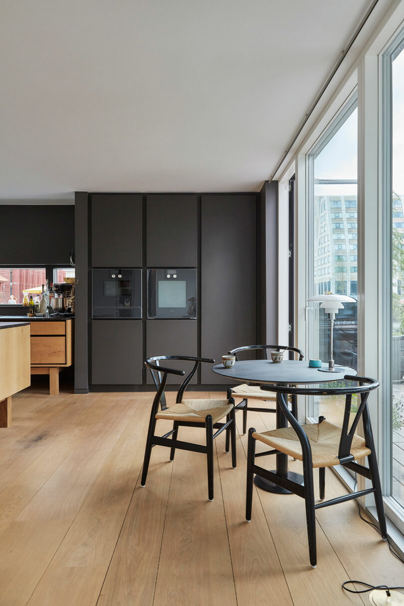 Modern kitchen with light wood flooring, black cabinetry, a small black dining table with two chairs, and large windows letting in natural light.