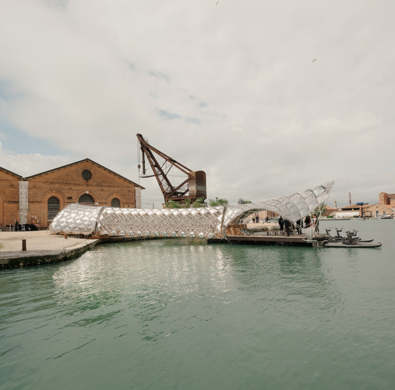 A modern, metallic, tunnel-like structure spans a canal beside brick industrial buildings and a large crane under a partly cloudy sky.
