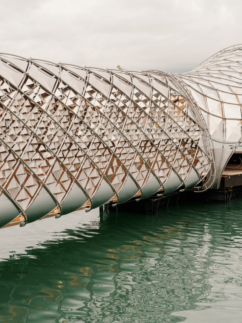A modern structure with a geometric metal and glass design extends over green water under a cloudy sky.