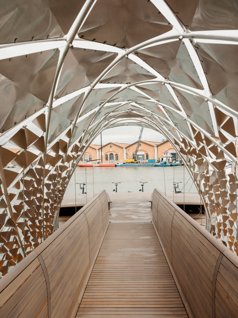 A wooden pedestrian bridge with a geometric metal canopy spans over water, leading toward industrial-style buildings in the background.
