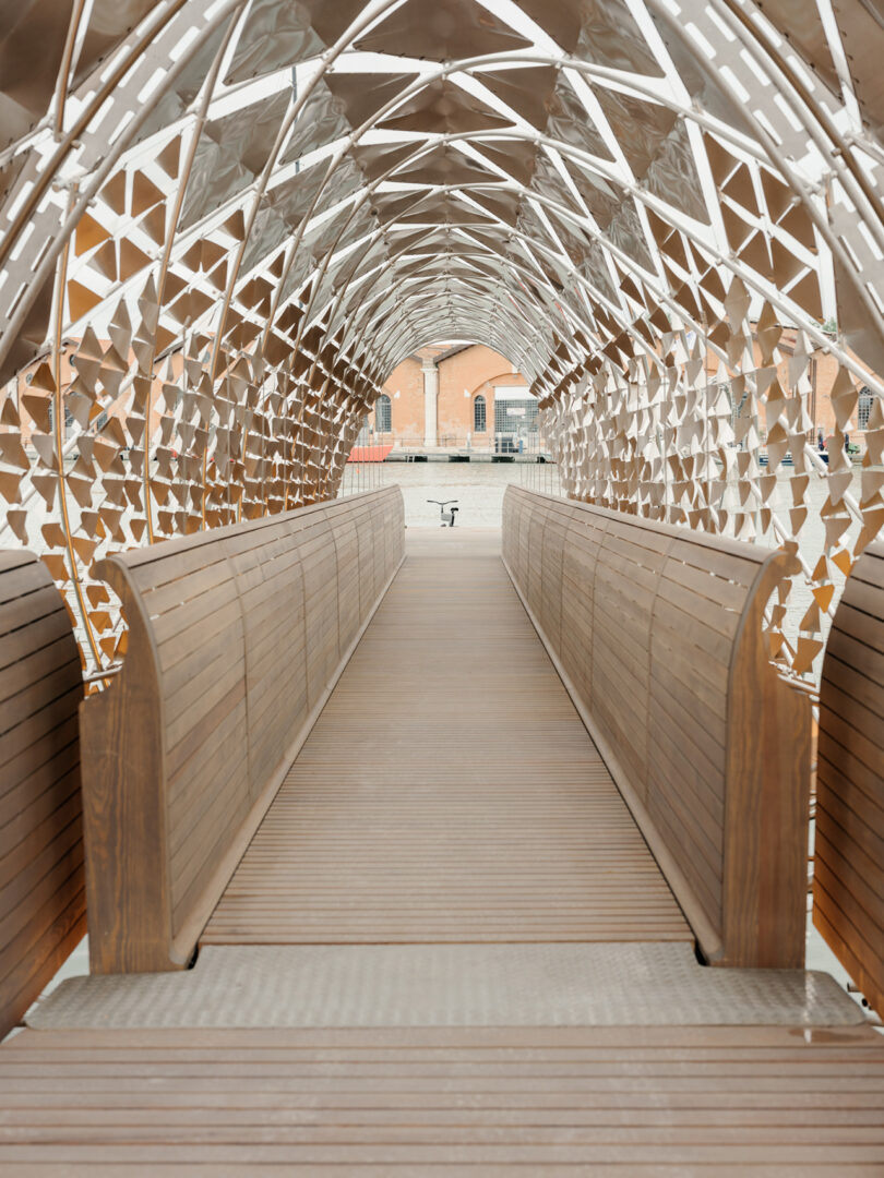 A wooden pedestrian bridge with curved sides and a geometric metal canopy leads to a building in the background.